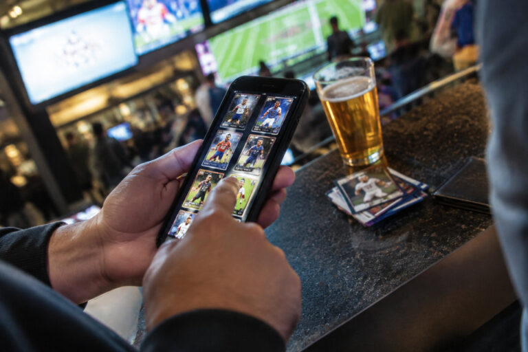 Collection de cartes numériques ou jeu d’argent : la frontière PrizePicks après les rachats instantanés Mains tenant un smartphone affichant des cartes de sport numériques dans un bar avec un verre de bière et des cartes réelles.