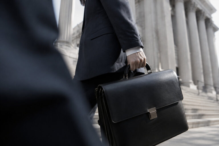Une personne en costume porte une mallette noire devant un bâtiment à colonnes de type tribunal.